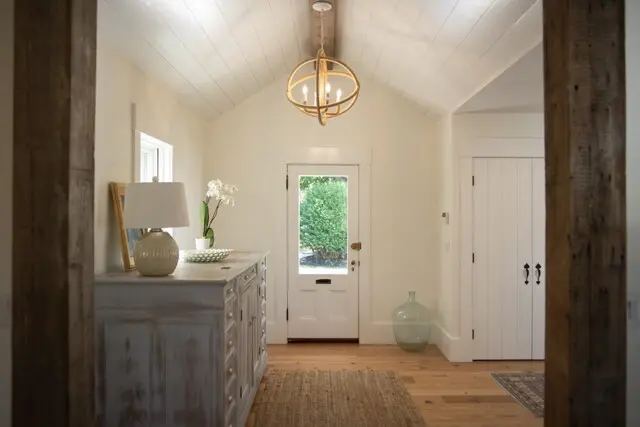 Cozy entryway with white paneled ceiling, a circular rope chandelier, and a distressed gray sideboard topped with a lamp and vase; door leads outside.