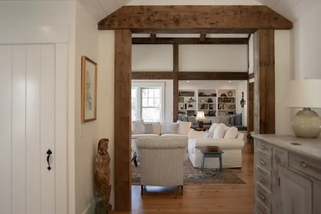 Bright living room seen through rustic wooden doorway with large timber beams; white sofas, striped chair, bookshelves, and warm natural light.