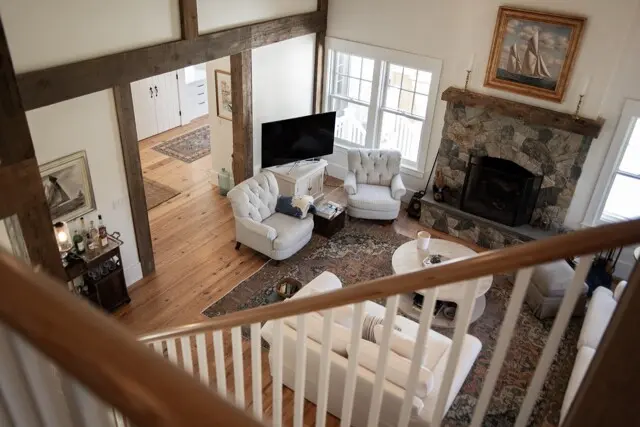 Cozy living room seen from a balcony: stone fireplace, large windows, white tufted chairs, sofa, TV on a console, and a patterned rug over wooden floors.