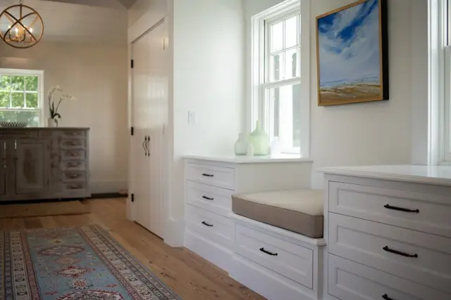 Bright white built-in window seat with drawers and beige cushion, green glass vases on ledge, a landscape painting, and light wood floors in a sunny, tidy room.