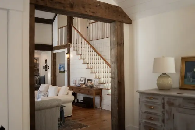 A cozy living space with a wooden framed doorway, white staircase, and cream furniture. A rustic console table, framed photos, and a distressed dresser with a lamp are visible.
