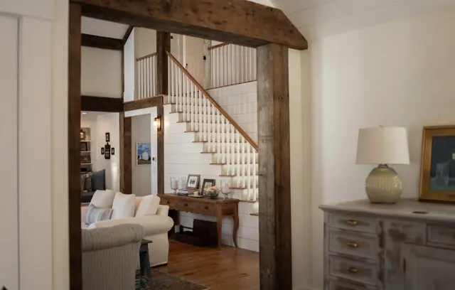 A cozy living space with a wooden framed doorway, white staircase, and cream furniture. A rustic console table, framed photos, and a distressed dresser with a lamp are visible.