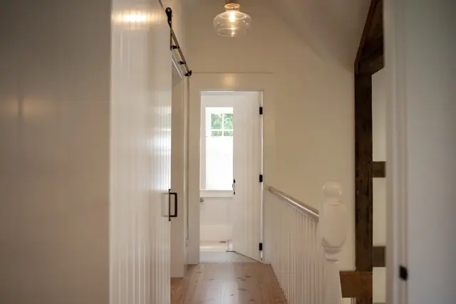 Bright, narrow hallway with white paneled walls, a wooden floor, and a white railing leading to a door at the end letting in natural light from a window. A glass ceiling light hangs overhead.