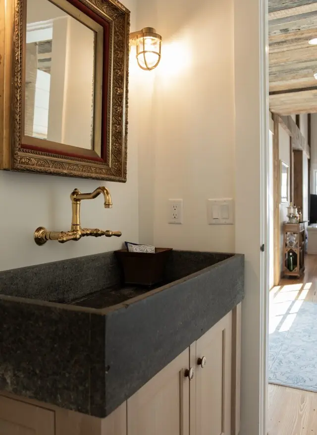A rustic bathroom sink with a dark stone countertop, brass wall faucet, an ornate framed mirror, and warm wall light; light switches and a wooden cabinet beneath.