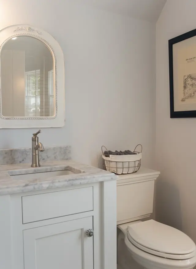 White bathroom with marble countertop, light gray walls, and a vintage arched mirror. A sink with brushed nickel faucet, a basket on the toilet tank, and framed art on the wall.