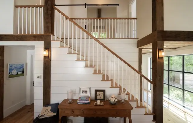 Bright two-story foyer with white shiplap walls, a wooden staircase and beams, a rustic console table, and large windows bringing in natural light.