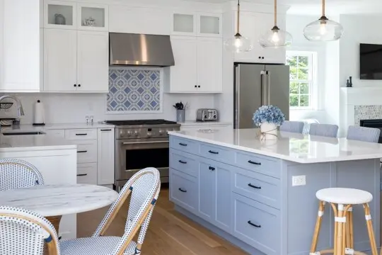 Bright white and light blue kitchen with a large island, stainless steel appliances, glass pendant lights, and a blue hydrangea centerpiece. Woven chairs nearby.