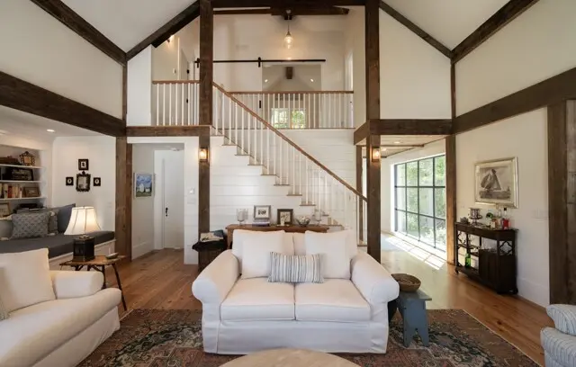 Bright open living room with white sofas, wooden beams, and a staircase leading to a loft. Large window wall, bookshelves, and a patterned rug.