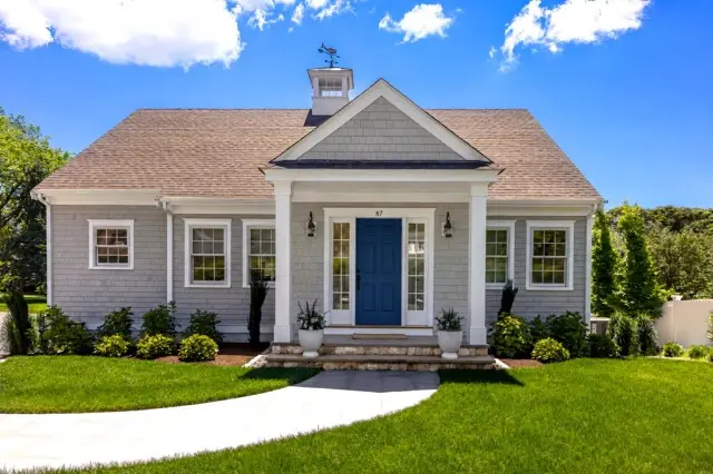 A charming single-story gray clapboard house with a blue front door, white trim, and a small covered porch. Bright green lawn and clear blue sky.