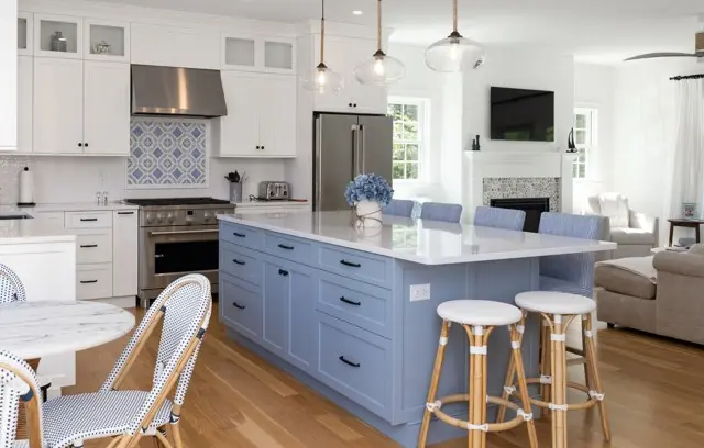Bright, airy kitchen with white upper cabinets, blue island, stainless steel appliances, blue patterned backsplash, and a living area with a fireplace and TV.