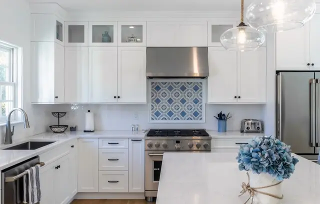 Bright white kitchen with glass-front cabinets, stainless range and hood, blue patterned tile behind stove, and a light marble island with blue hydrangea centerpiece.