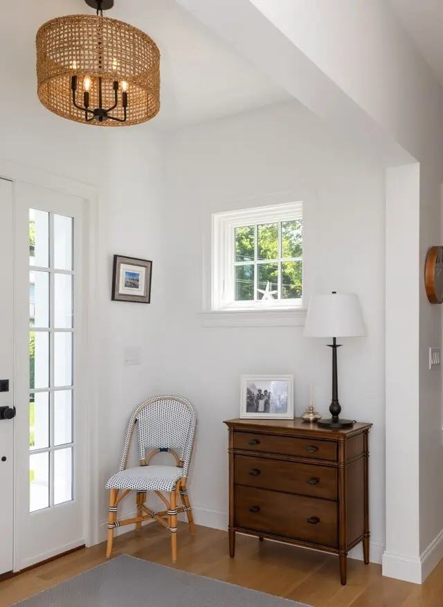 Bright entryway with white walls, a woven pendant light, a wooden dresser topped with a lamp and photos, a patterned chair, and a small window letting in greenery.