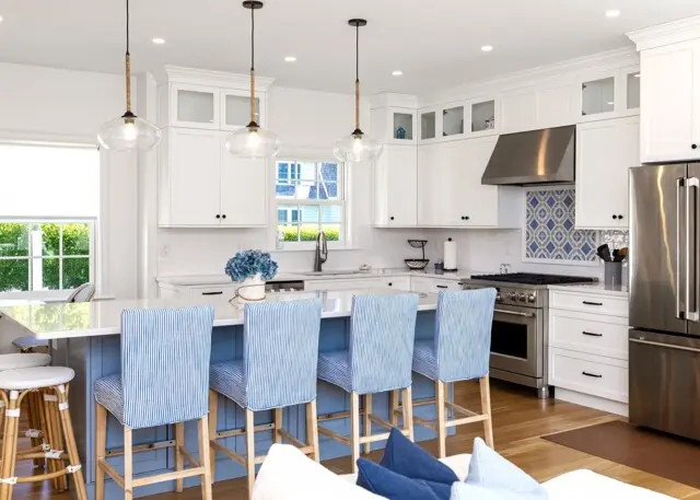 Bright white kitchen with blue island, four striped blue bar chairs, stainless steel fridge and oven, glass pendant lights, and a patterned tile backsplash.