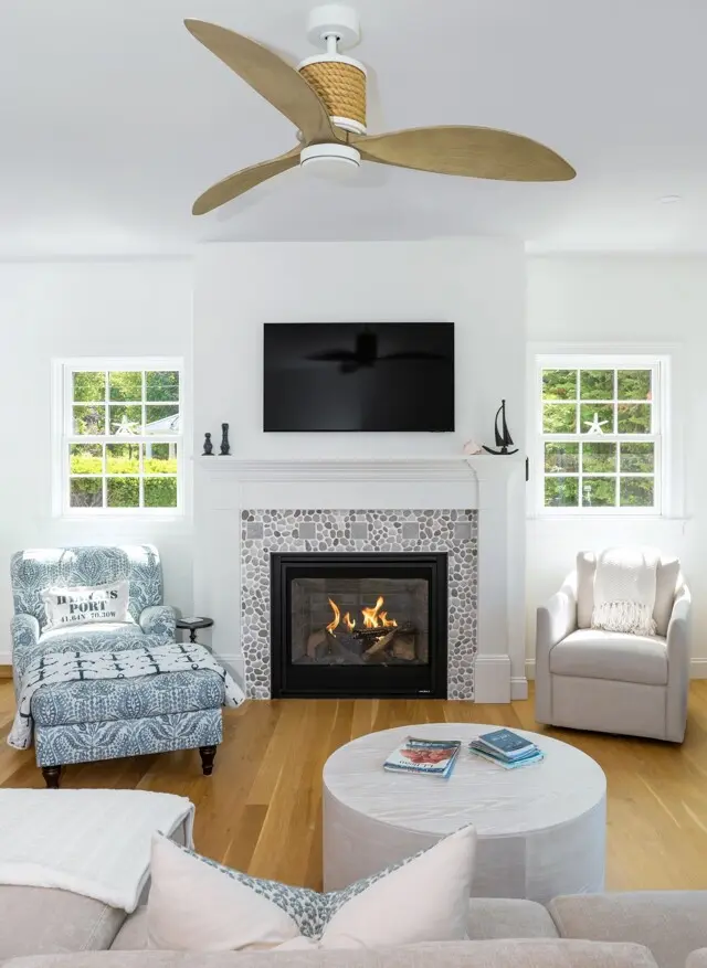 Bright living room with a modern white fireplace, mosaic hearth, mounted TV, and a ceiling fan. Light blue pattern chair, white armchair, round coffee table, and sunny windows.