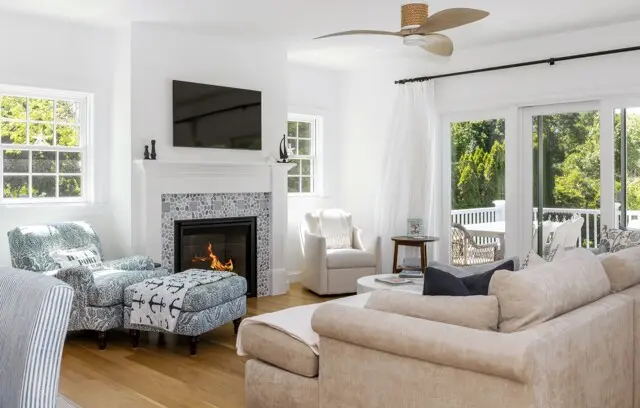 Bright living room with a gas fireplace, mounted TV above, and white walls. Light seating includes a beige sectional and patterned chairs; large windows and sliding doors open to a sunny outdoor deck.