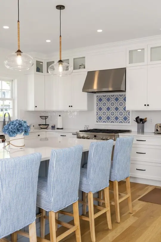 Bright white kitchen with a blue tiled backsplash, stainless range, and island seating; light blue striped bar stools and glass pendant lights.