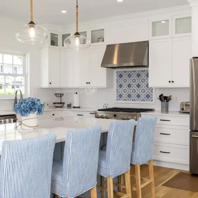 Bright white kitchen with a blue tiled backsplash, stainless range, and island seating; light blue striped bar stools and glass pendant lights.