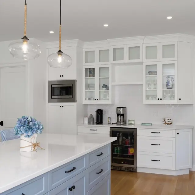 Bright white kitchen with glass-front cabinets, stainless micro, coffee station, built-in wine fridge, and a large island with blue hydrangea centerpiece.