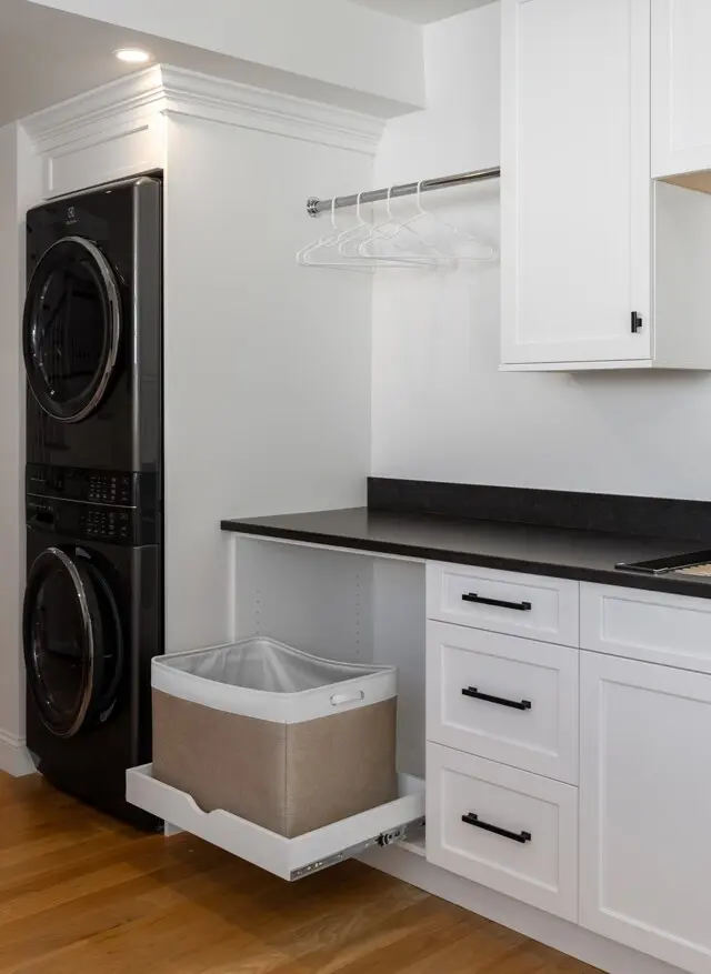 Modern laundry area with stacked front-load washer/dryer, white cabinetry, black countertop, hanging rod with white hangers, and a pull-out woven laundry basket.