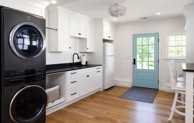 Bright, modern laundry room with white cabinets, black countertops, stacked washer/dryer, and a light blue door; wood floor, black faucet, and a small rug.