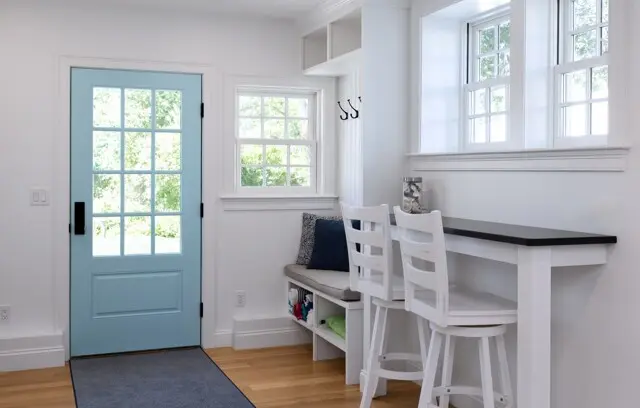 Entryway with a light blue door, white walls, and wooden floor. Built-in seating with cushions, a black-counter bar with white stools, and large windows letting in sunlight.