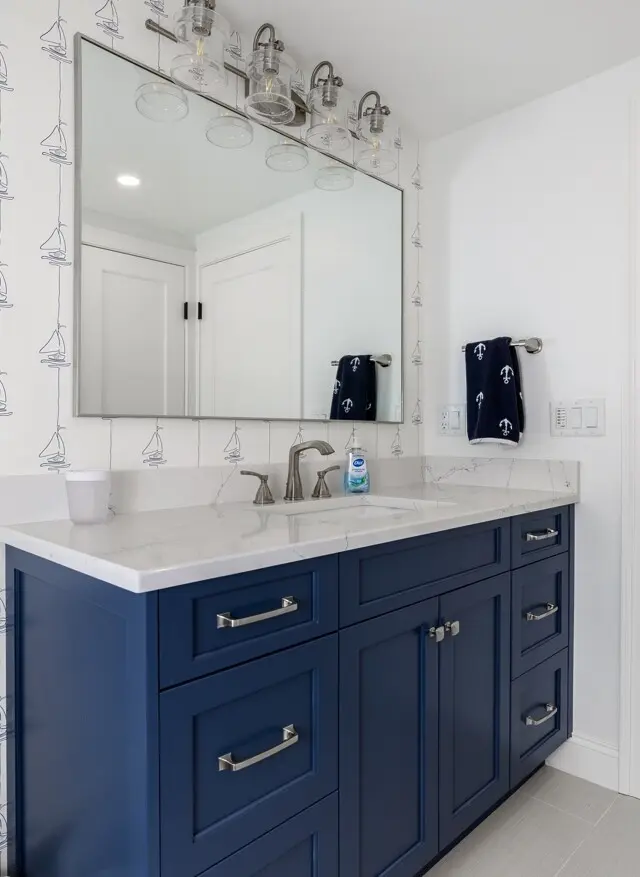 Navy blue bathroom vanity with a white marble countertop and silver hardware, large rectangular mirror, wall with sailboat wallpaper, and navy towels on a chrome bar.