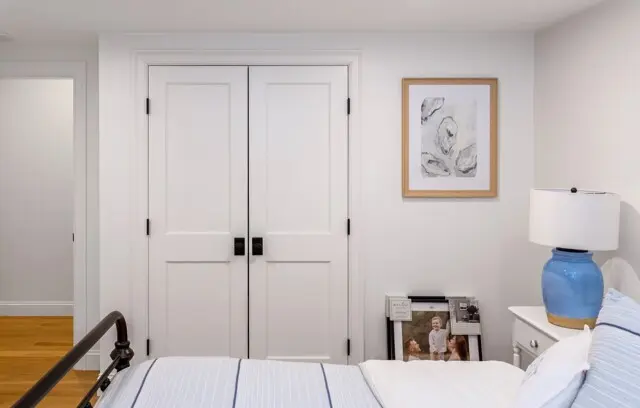 Bright bedroom with white double doors, a framed abstract print, a blue ceramic lamp on a white nightstand, and a nearby stack of magazines by the bed.