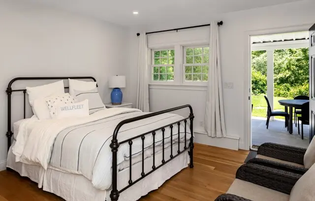 Bright bedroom with a dark metal bed, white bedding, blue lamp, and light gray curtains. A window overlooks greenery; a door opens to a patio. Oak floor.
