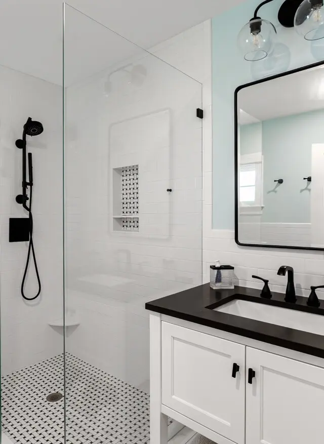 Modern white bathroom with a glass shower enclosure, black hardware, and a black countertop. White cabinetry, black faucet, and a large framed mirror.
