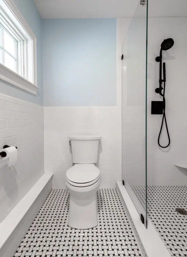 Light bathroom with light blue upper walls, white tiled wainscoting, and a toilet. A glass shower door and black shower fixture are on the right. Window on left.