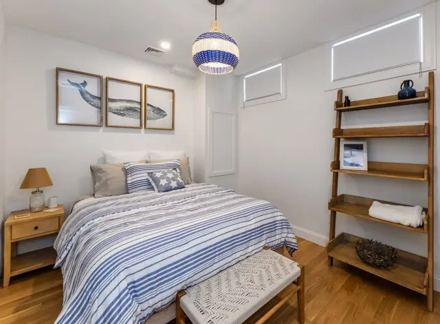 Cozy coastal bedroom with a striped blue-and-white bed, wooden nightstand, woven pendant light, sea-themed art above the bed, and a wooden ladder shelf.