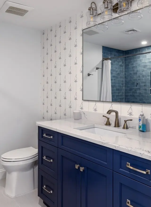 Blue vanity with white marble countertop, brass faucet, and navy cabinets; white tiled wall with sailboat pattern, large mirror, and blue tiled shower area. Toilet nearby.