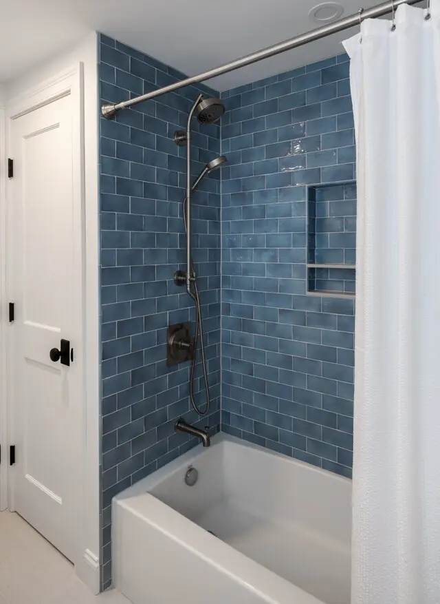 Blue subway-tiled shower walls with a built-in niche, a dark metal handheld shower, a fixed shower head, and a white bathtub. White shower curtain on a rod. White door nearby.