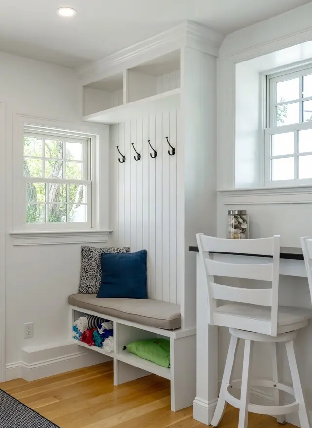 Bright white entry nook with a built-in bench, beadboard wall, and black coat hooks. Windowed light, cushions, open cubbies with colorful items, and a small desk with white chairs.