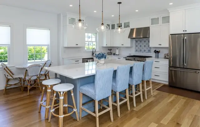 Bright open kitchen with white cabinetry, stainless steel fridge and range, light wood floors, and a blue-gray island with four striped blue stools. Glass pendant lights. Coastal style.