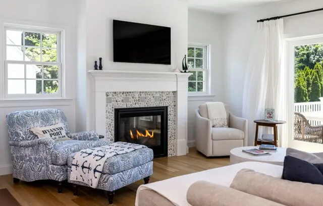 Bright living room with a white fireplace, pebble mosaic hearth, wall-mounted TV, blue patterned chaise, cream chair, and large windows opening to a sunny outdoor space.