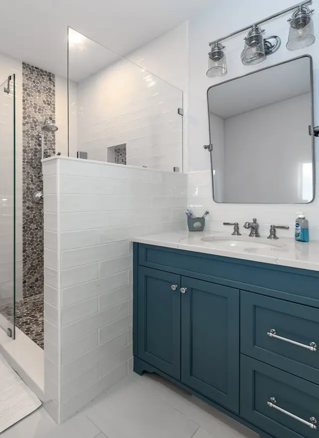 Modern bathroom with a glass shower enclosure, white subway tiled walls, and a teal vanity with a white countertop. Large mirror and three-bulb metallic light fixture.