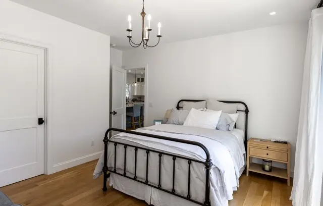 Bright, minimalist bedroom with white walls, a dark metal bed frame, white bedding, light wood floors, and a small wooden nightstand. A chandelier hangs from the ceiling.