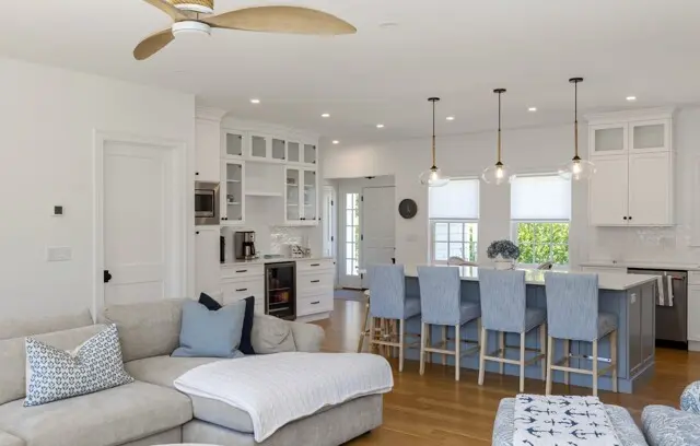 Bright, open kitchen and living area with white cabinets, light blue island stools, hardwood floors, ceiling fan, and large windows letting in natural light.