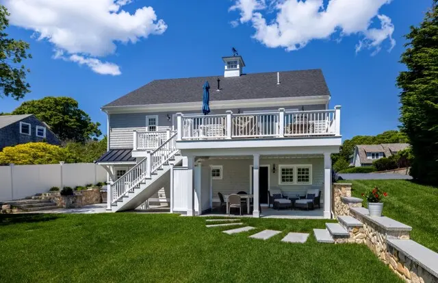 Two-story coastal-style home with a white deck and stairs, gray siding, outdoor seating, green lawn, stone accents, and blue sky.