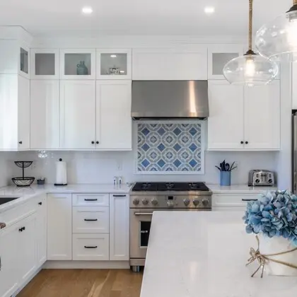 Bright white kitchen with upper cabinets, stainless range, geometric blue tiled backsplash, and a large island topped in white; blue hydrangea bouquet on island.