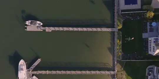 Aerial view of a marina with a long wooden dock extending from the shoreline, a white boat at the end, and houses with green lawns on the right.
